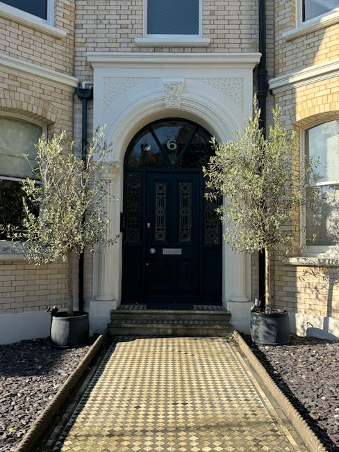 The image depicts the front entrance of a residential property with a classic architectural style, featuring a black front door with decorative glass panels and a rectangular letterbox. The door is framed by an elegant cream-colored stone archway with intricate carvings and a keystone at the top. Flanking the doorway are two potted olive trees with silvery-green foliage, planted in large black pots. The pathway leading to the door is a patterned mosaic tile with a diamond design in beige and black, bordered by narrow wooden edges. The surrounding garden area displays a neat, low-maintenance appearance with dark mulch beds on either side, contrasting with the light brick exterior of the house. The overall scene captures a well-maintained and tidy entrance that highlights quality garden and landscape features. This is an example of garden maintenance and landscaping work by Gardeners Acton, emphasizing a tidy and inviting outdoor entrance consistent with the Acton Park front garden makeover case study.