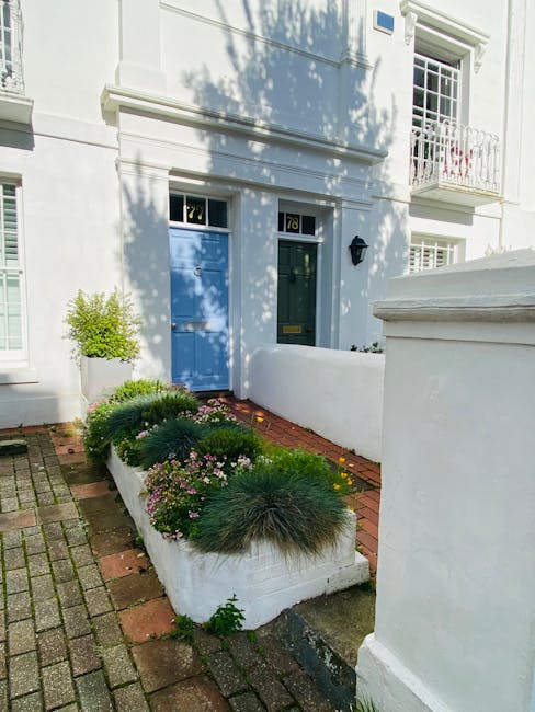 The image depicts the front entrance of a residential property with a classic architectural style, featuring a black front door with decorative glass panels and a rectangular letterbox. The door is framed by an elegant cream-colored stone archway with intricate carvings and a keystone at the top. Flanking the doorway are two potted olive trees with silvery-green foliage, planted in large black pots. The pathway leading to the door is a patterned mosaic tile with a diamond design in beige and black, bordered by narrow wooden edges. The surrounding garden area displays a neat, low-maintenance appearance with dark mulch beds on either side, contrasting with the light brick exterior of the house. The overall scene captures a well-maintained and tidy entrance that highlights quality garden and landscape features. This is an example of garden maintenance and landscaping work by Gardeners Acton, emphasizing a tidy and inviting outdoor entrance consistent with the Acton Park front garden makeover case study.