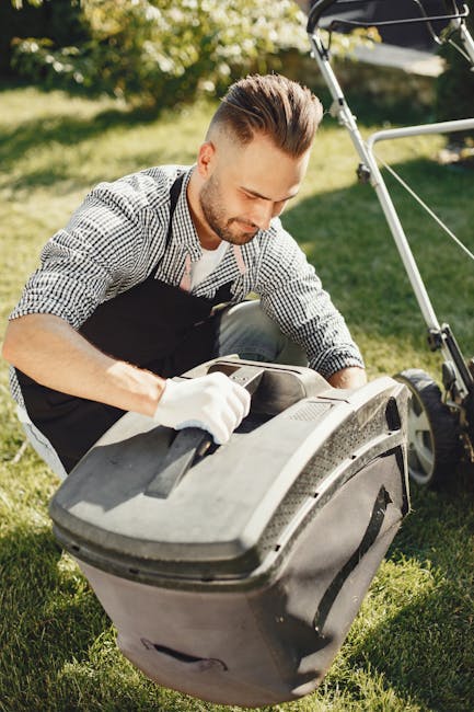 A man with styled brown hair and a short beard, dressed in a checkered shirt and black apron, is kneeling on a lush green lawn while attending to garden maintenance. He is wearing white gloves and appears focused as he opens a large grey garden waste bin, which is positioned on the neatly trimmed grass. The garden surrounding him features well-maintained lawns, garden beds with dark, moist soil, and a hedge in the background. To his right, part of a lawnmower with a handle and a grass collection bag is visible, indicating ongoing lawn care work. Soft sunlight illuminates the scene, highlighting the textures of the grass, leaves, and the man's clothing. This setting reflects professional gardening and landscaping activities, emphasizing garden upkeep in a typical outdoor environment. The image naturally promotes garden maintenance services offered by Gardeners Acton, aligning with themes of lawn care, hedge trimming, and outdoor upkeep.