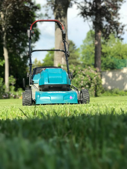A man with styled brown hair and a short beard, dressed in a checkered shirt and black apron, is kneeling on a lush green lawn while attending to garden maintenance. He is wearing white gloves and appears focused as he opens a large grey garden waste bin, which is positioned on the neatly trimmed grass. The garden surrounding him features well-maintained lawns, garden beds with dark, moist soil, and a hedge in the background. To his right, part of a lawnmower with a handle and a grass collection bag is visible, indicating ongoing lawn care work. Soft sunlight illuminates the scene, highlighting the textures of the grass, leaves, and the man's clothing. This setting reflects professional gardening and landscaping activities, emphasizing garden upkeep in a typical outdoor environment. The image naturally promotes garden maintenance services offered by Gardeners Acton, aligning with themes of lawn care, hedge trimming, and outdoor upkeep.